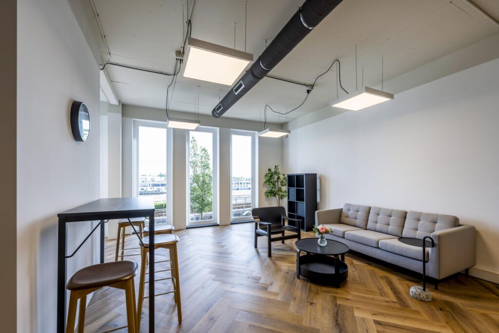 Moderne wachtruimte aan de De Ruijterkade met een grijze bank, hoge tafel met krukken en grote ramen met uitzicht op de stad.